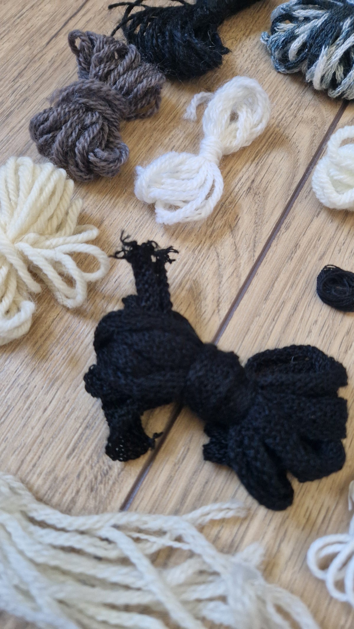 A close-up of a black knotted yarn bundle placed on a light wood surface, surrounded by other skeins of yarn in cream, white, gray, and black.