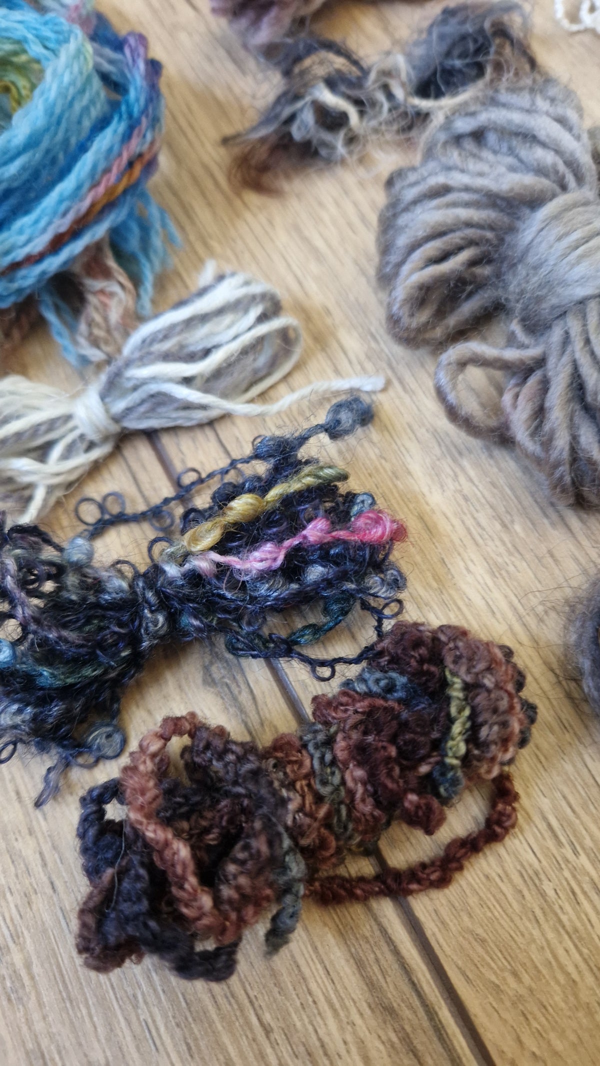 Several skeins of textured, curly yarn in various colors, including brown, gray, blue, and black, arranged on a light-colored wooden surface.