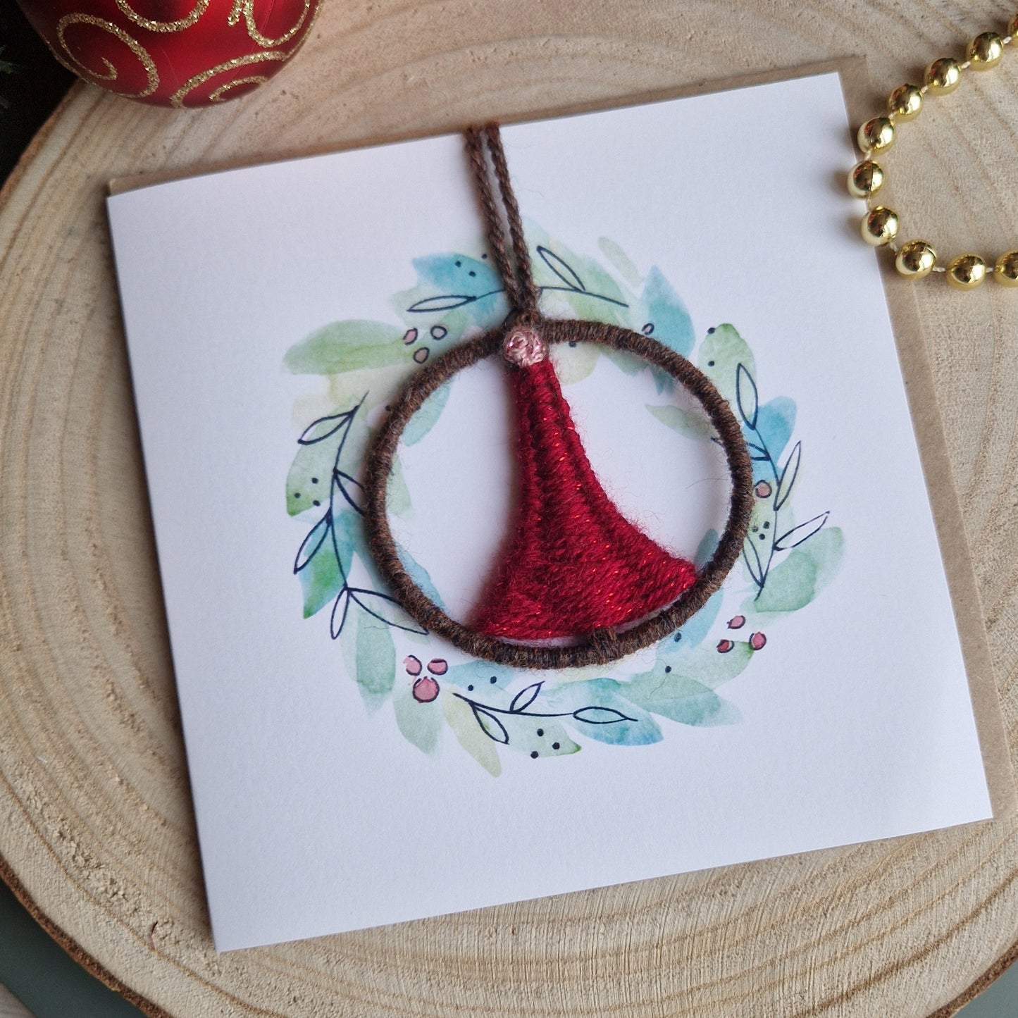 Card with a red and brown handwoven ornament on a wooden surface with Christmas ornaments.