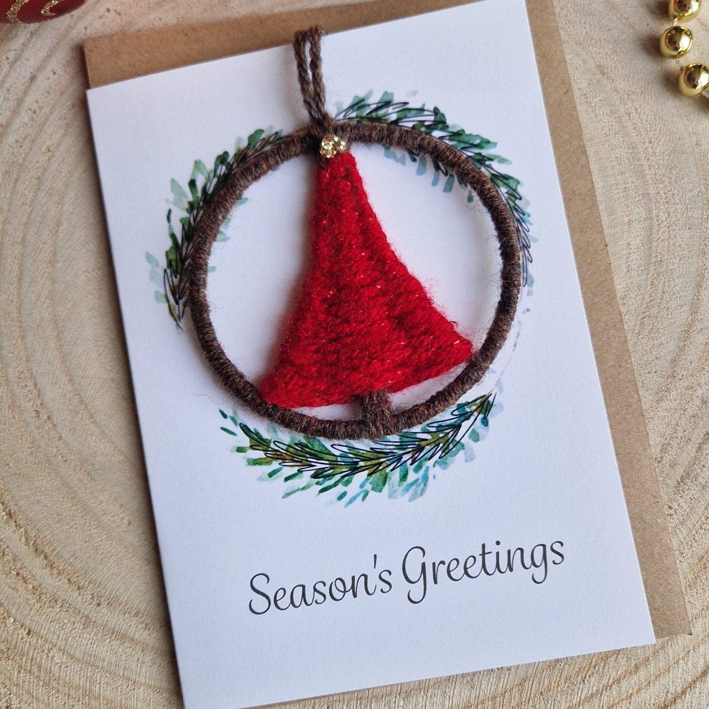 Greeting card with red Christmas Tree shaped ornament and greenery wreath design on a wooden surface with Christmas ornaments.