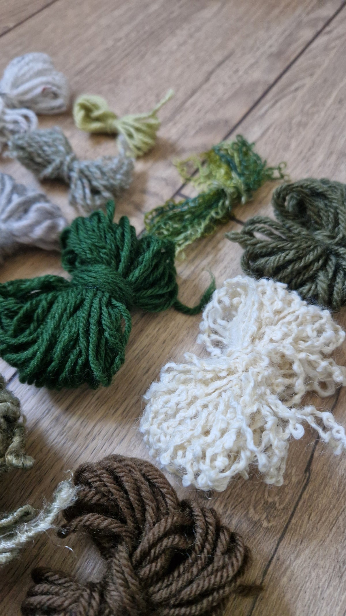 A variety of knotted yarn skeins in different colors, including forest green, cream, brown, and olive, arranged on a light wood floor.