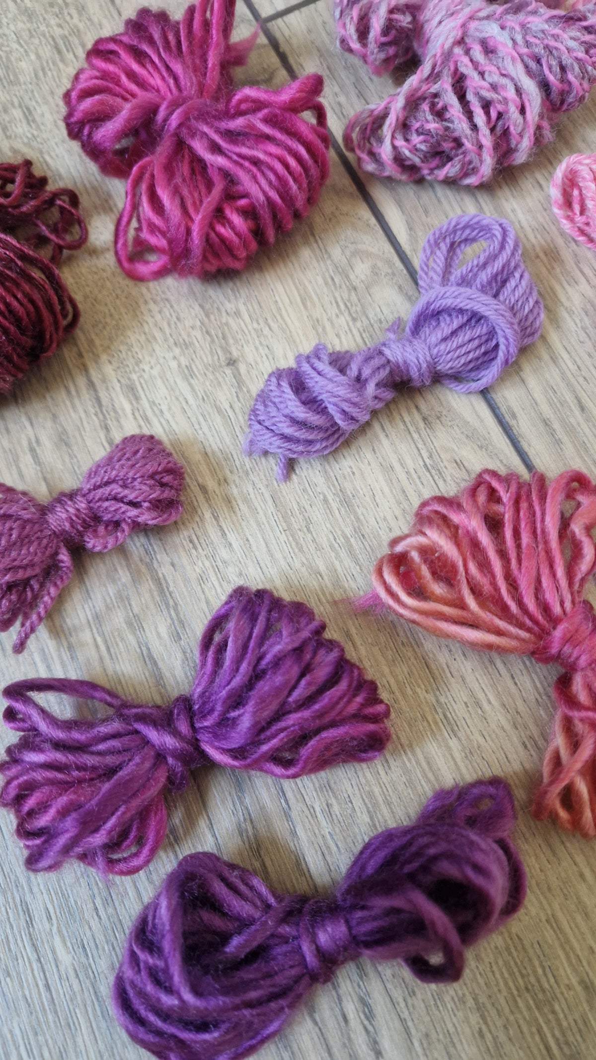 Several small bundles of knotted yarn in shades of pink, purple, and burgundy arranged on a light wood surface.