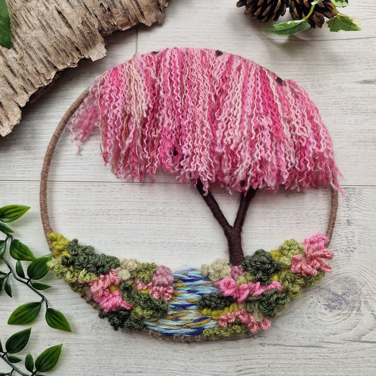 Cherry blossom tree made of yarn in a hoop frame. The base has a blue stream with green and pink yarns either side to depict vegetation.