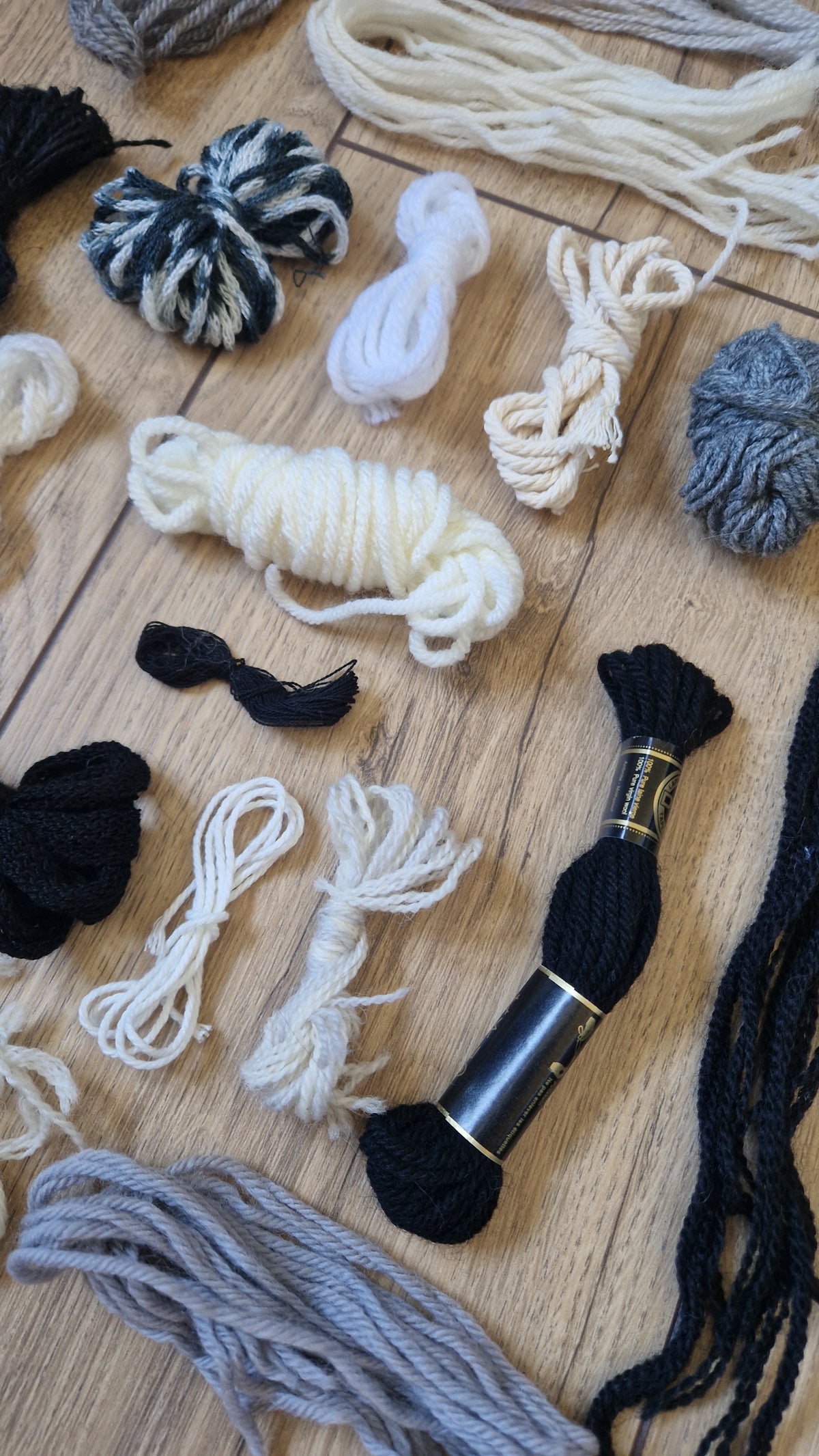 An assortment of yarn skeins and thread bundles in black, white, and gray laid out on a light wood floor. A black spool of yarn with gold lettering is visible near the bottom right.