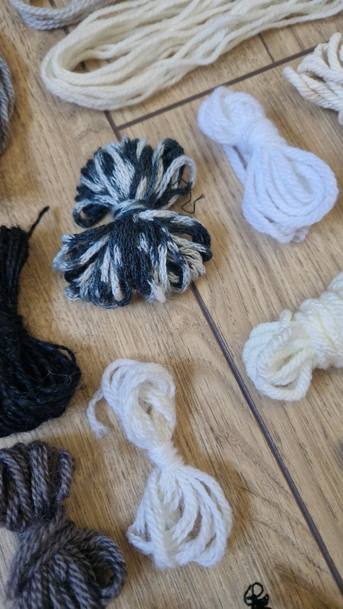 Several skeins of yarn in different colors and patterns arranged on a light wood floor, including cream, white, black, gray, and marled yarn.