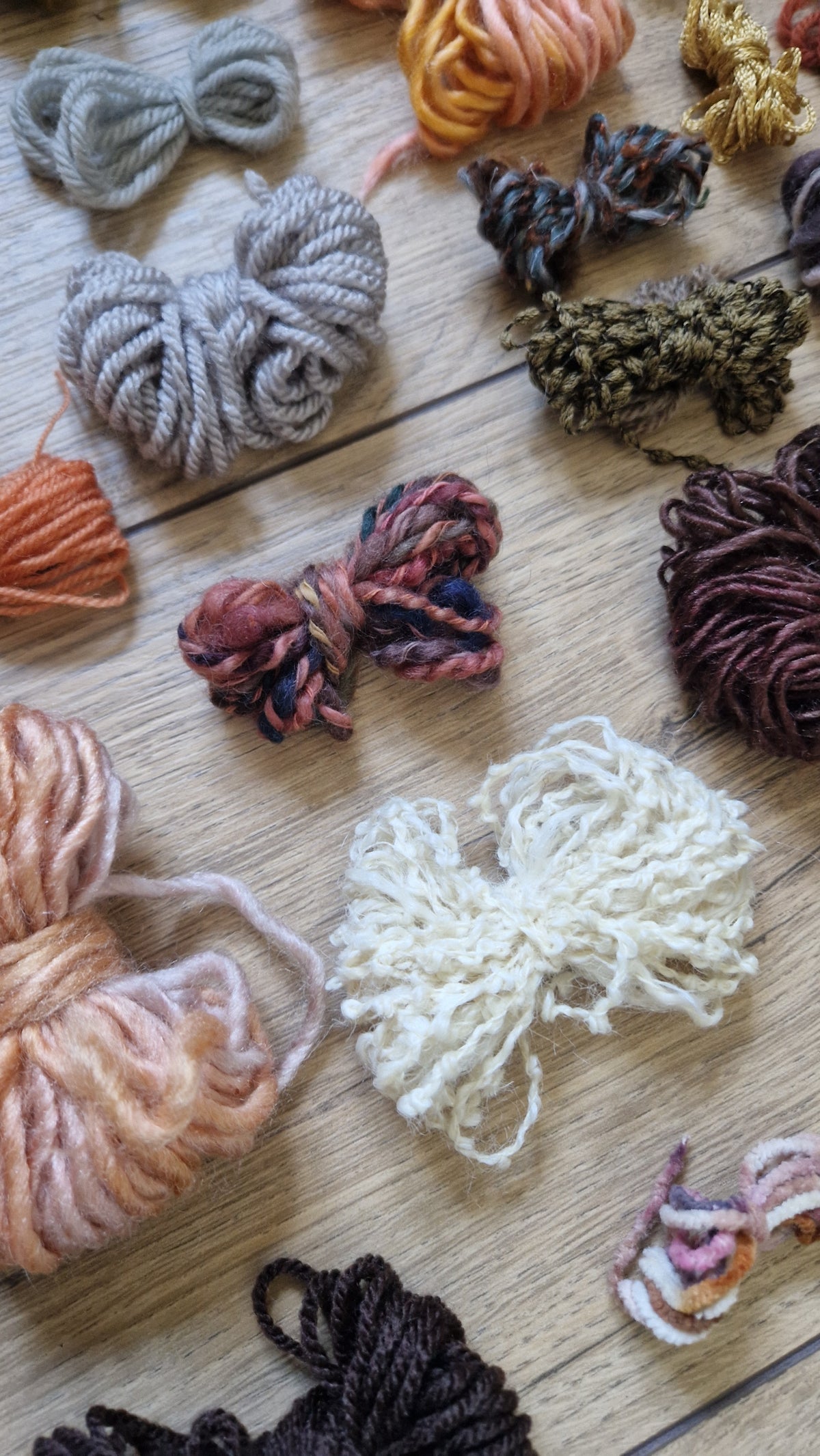An assortment of colourful yarn skeins and bundles arranged on a light wood surface, including shades of cream, orange and brown.