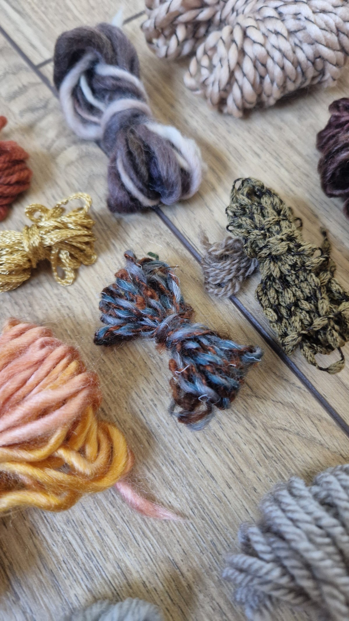 A close-up flat lay of multiple skeins of yarn in assorted colors and textures arranged on a light wood surface. Visible hues include cream, taupe, gray, blue-gray, rust orange, mustard yellow, metallic gold, olive green, and charcoal.