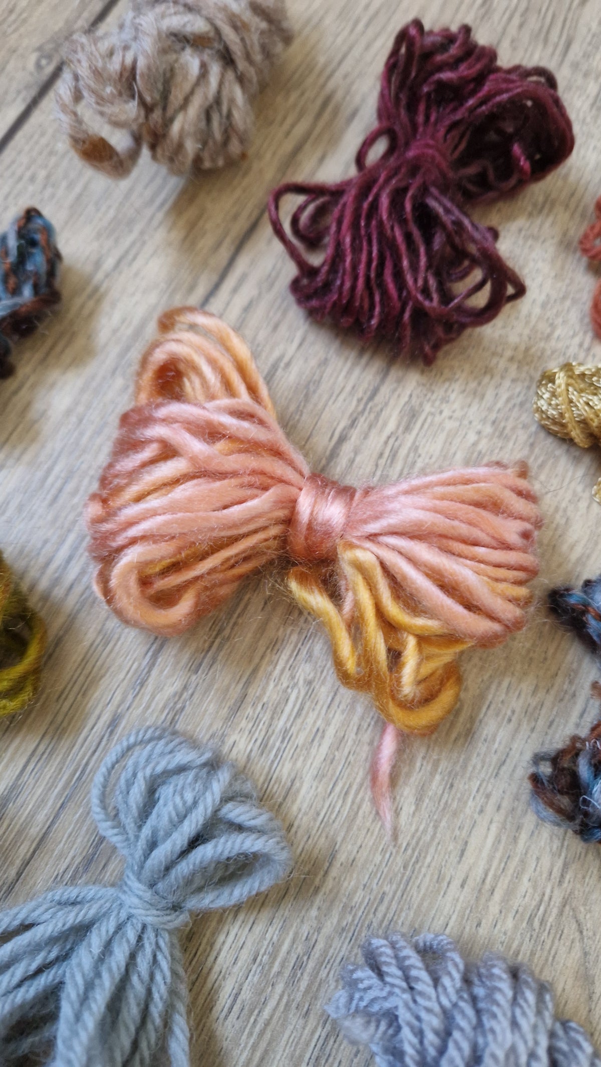 A styled flat-lay of various skeins of yarn in different colors, featuring a prominent peach and gold gradient skein in the center, surrounded by other skeins on a light wood background.