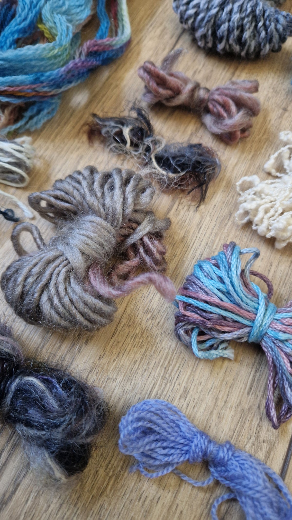 Multiple small bundles of twisted yarn in assorted colors and textures arranged on a light wood surface. The yarn includes shades of blue, gray, brown, cream, pink, and purple.