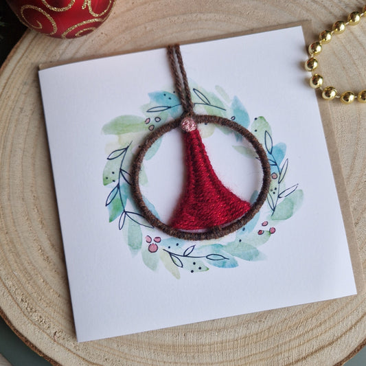 Card with a red and brown handwoven ornament on a wooden surface with Christmas ornaments.