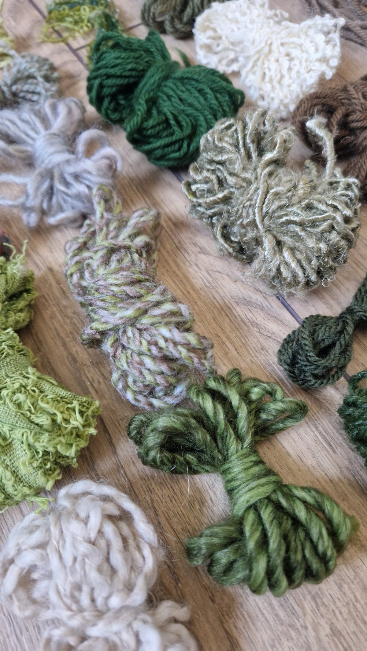 A variety of textured yarn skeins and bales in various shades of green, cream, and brown, arranged on a light wooden surface.