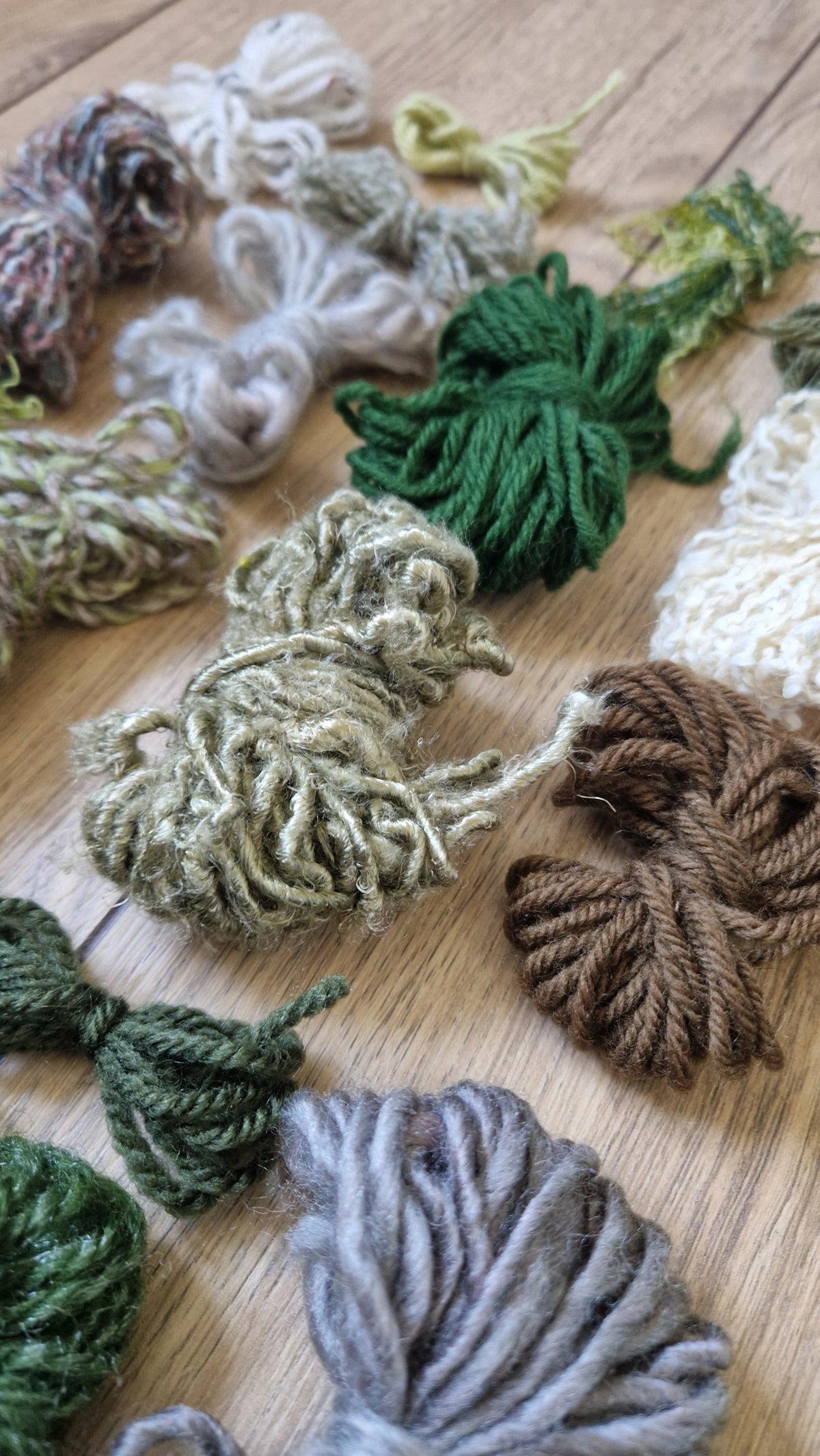 Multiple skeins of textured yarn in assorted earthy colors including green, brown, beige, gray, and cream, arranged on a light wood surface.