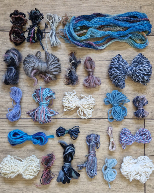 A collection of yarn skeins arranged in rows on a light wood floor. The yarns display a mix of colours, including various shades of blue, purple, grey, and cream.