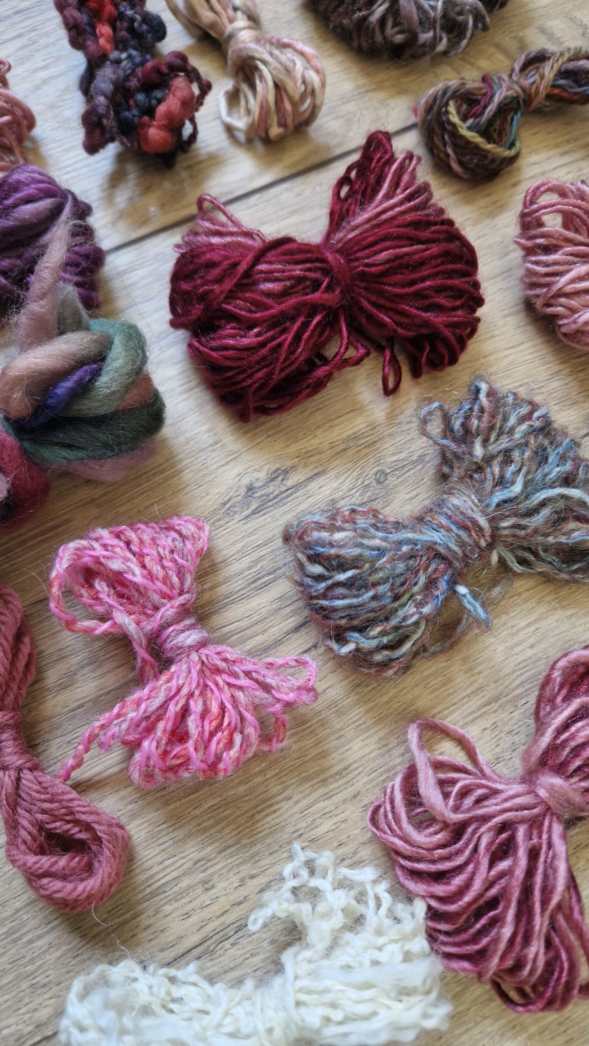 An assortment of colorful yarn skeins and bundles arranged on a light wooden surface, including shades of burgundy, pink, mauve, cream, and brown.