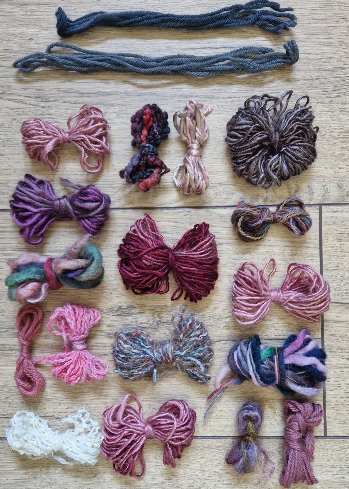 A collection of colourful yarn skeins and bundles arranged on a light wood floor. The yarns come in a variety of colors including pink, purple, grey, and white, with two darker gray yarn skeins laid out at the top.
