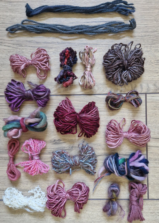 A collection of colourful yarn skeins and bundles arranged on a light wood floor. The yarns come in a variety of colors including pink, purple, grey, and white, with two darker gray yarn skeins laid out at the top.