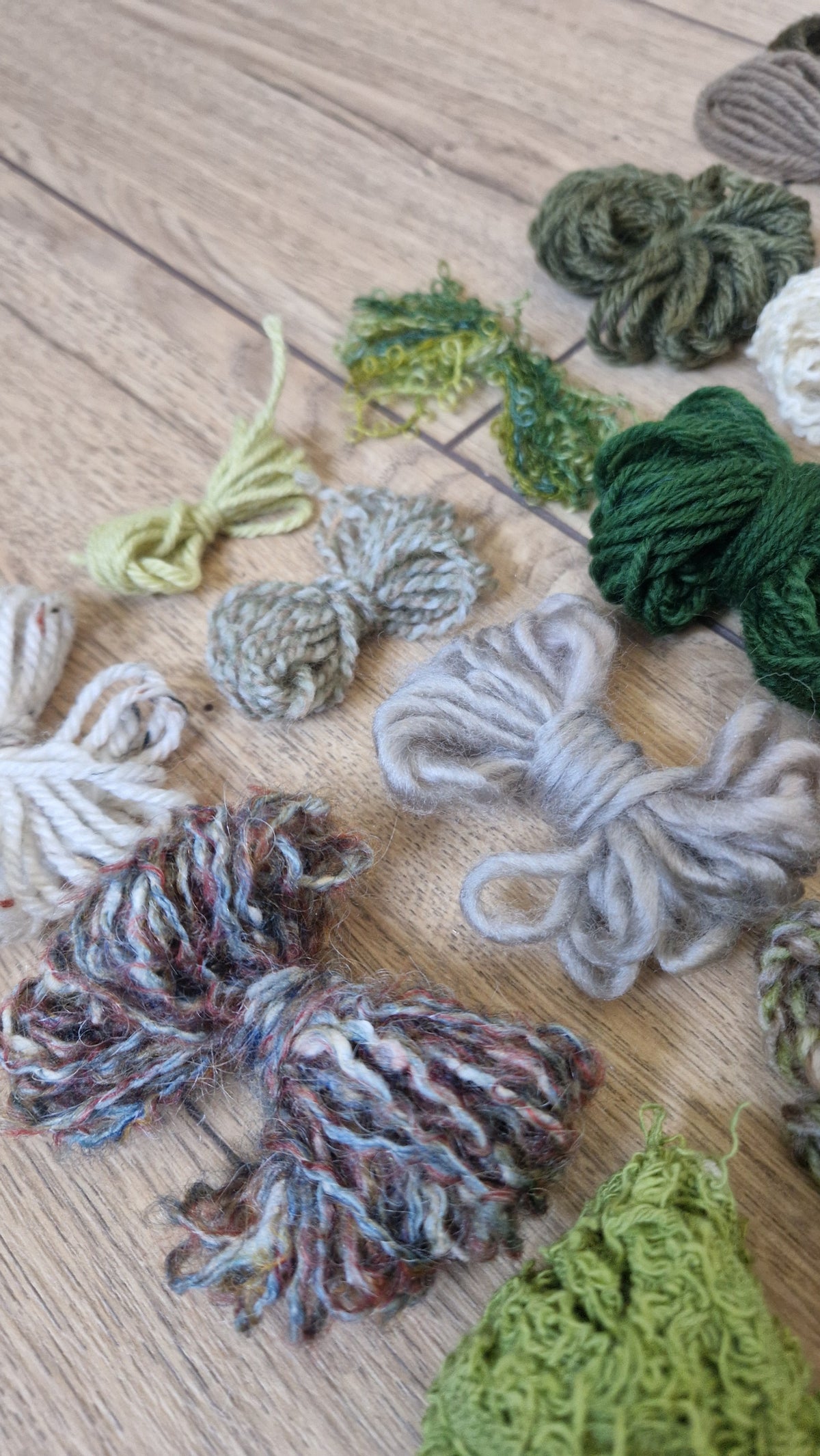 Several loose bundles of variegated yarn and a knitted fabric swatch arranged on a light wood floor. The yarns show mixed natural fiber colors including olive green, cream, gray, blue, and rust.