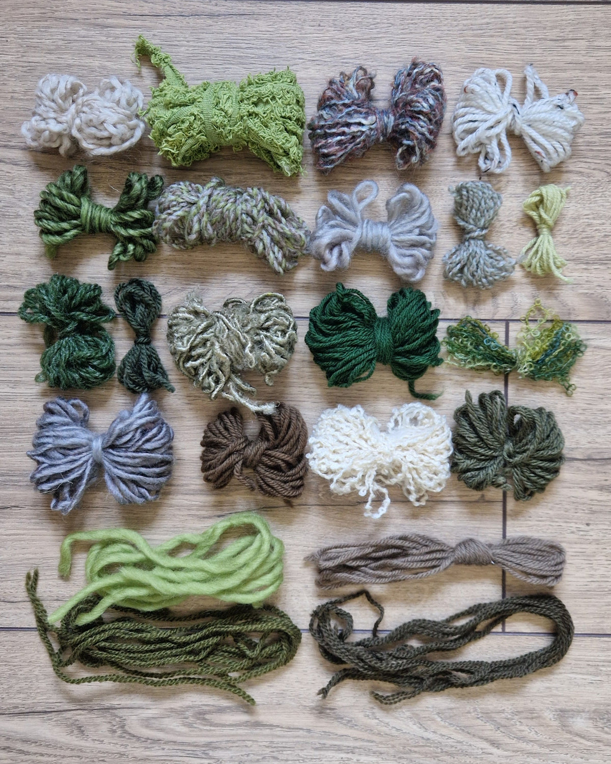 A flat-lay arrangement of various skeins and bundles of yarn in different colors and textures, organized in rows on a light brown wooden floor.