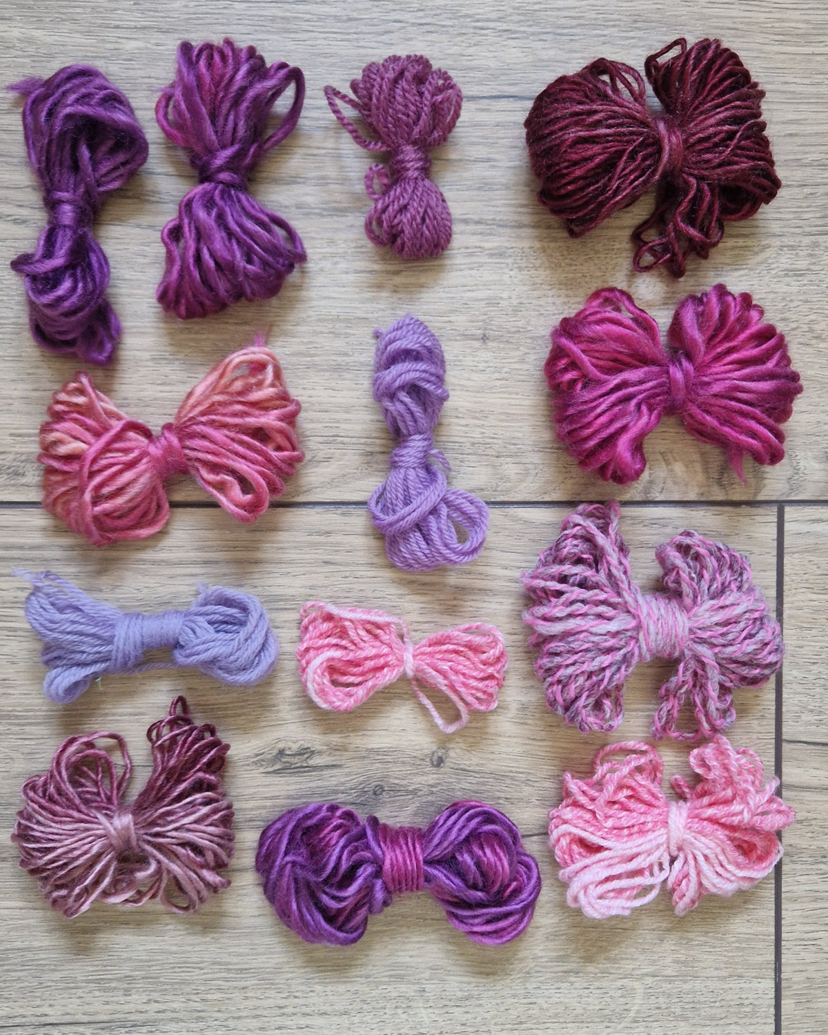 Skeins of variegated yarn in shades of purple, pink, and magenta, arranged in a grid on a light brown wood plank background.