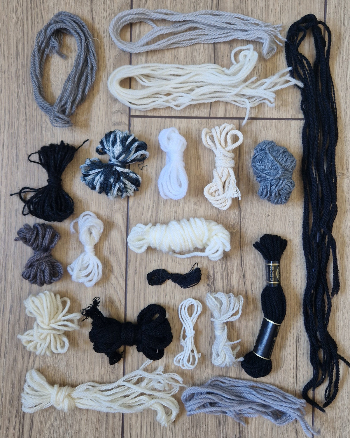 An assortment of yarn and embroidery floss skeins and bundles in black, white, gray, and beige, neatly arranged on a light brown wooden background.