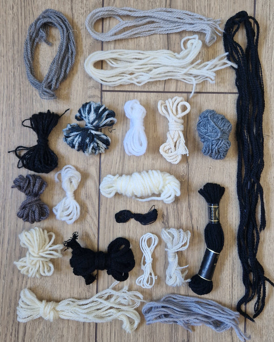An assortment of yarn and embroidery floss skeins and bundles in black, white, gray, and beige, neatly arranged on a light brown wooden background.