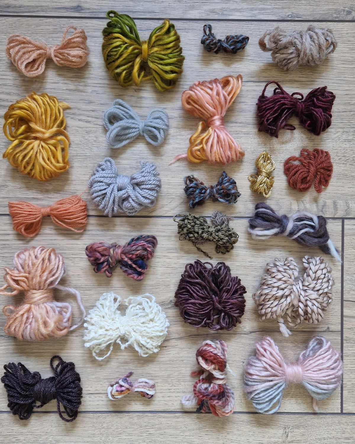 A flat-lay arrangement of various colorful yarn skeins and small fabric bows on a light wood floor. The collection features a wide variety of colors including mustard yellow, olive green, dusty pink, light blue, rust red, cream, and black.